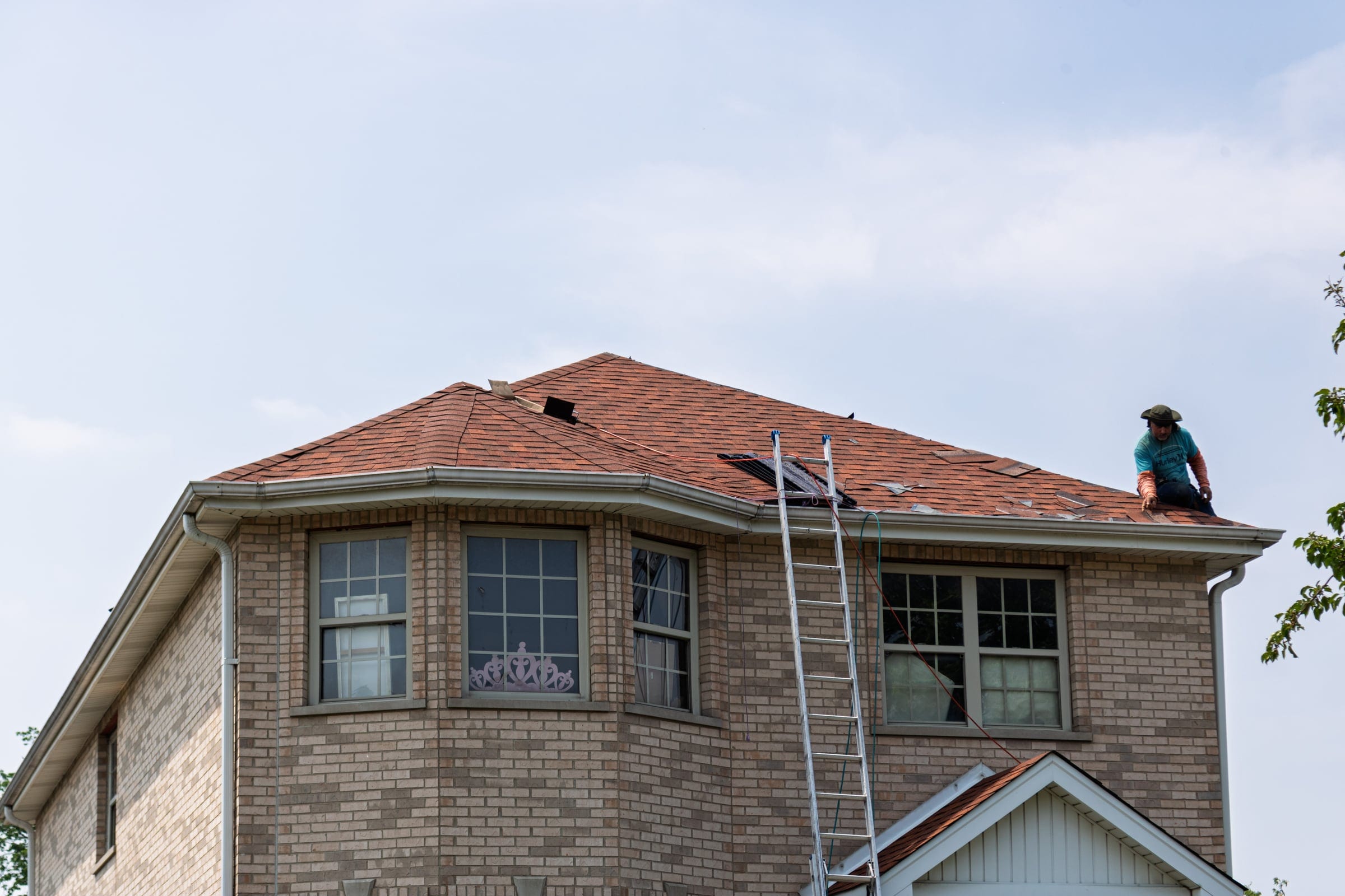A roofer at the eave of a brick home during a tear-off, with a ladder against the gutter